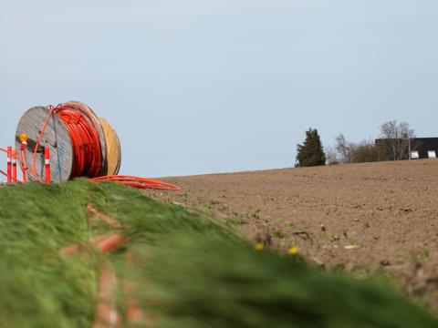 Große Holzkabeltrommel mit orangem Verbundrohr seitlich auf einem Grünsteifen neben einem Acker vorbereitet zur Erdverlegung. Einfamilienhaus im Hintergrund.