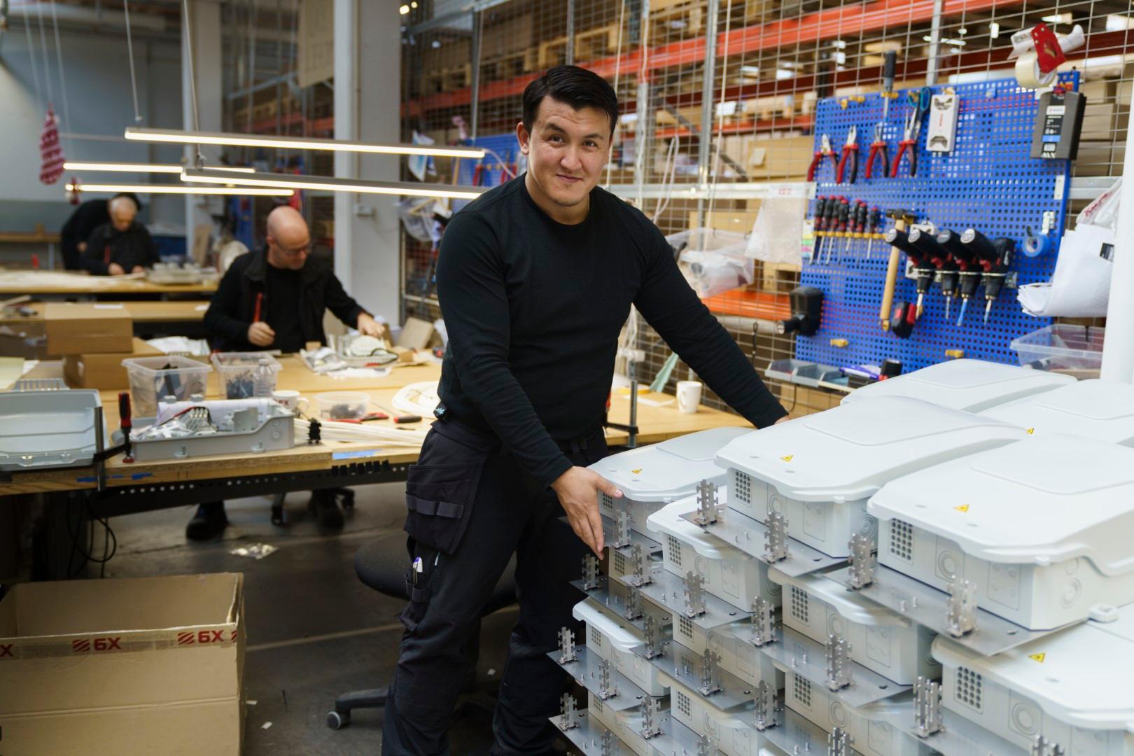 Produktionsmitarbeiter verpackt konfektionierte BUDI-Boxen. Im Hintergrund sitzen zwei Mitarbeiter an ihrem Arbeitstisch und montieren Spleißboxen.