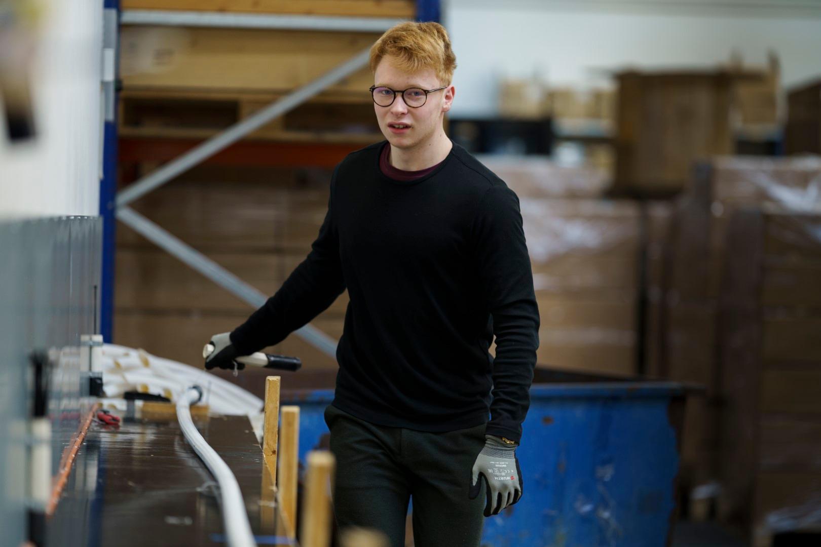 Ung mand med rødt hår og briller arbejder ved en produktionslinje med fiberrør i Alcadons produktionsafdeling. Han bærer handsker og sort trøje og kigger mod kameraet.