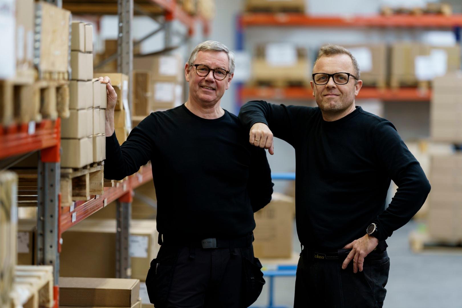 Zwei Lagermitarbeiter stehen am Hochregallager und lächeln in die Kamera.