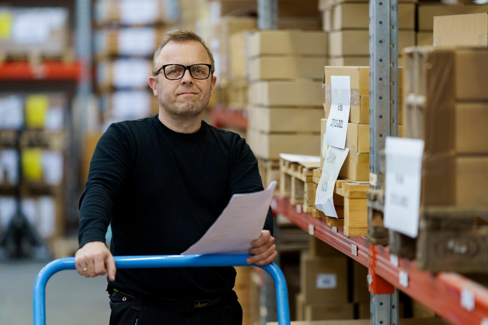 Lagermitarbeiter geht mit Packliste und Platformwagen durchs Hochregallager